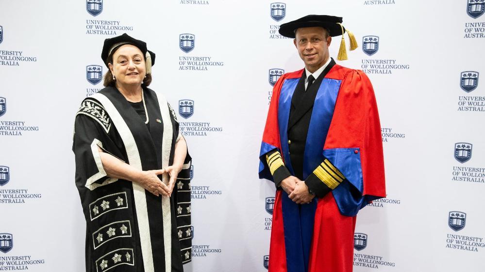 Vice Admiral Michael Noonan, wearing a red graduation gown, in front of a UOW media wall ahead. To his left is UOW Vice-Chancellor Professor Patricia M Davidson, in black graduation gowns and cap. Photo: Paul Jones