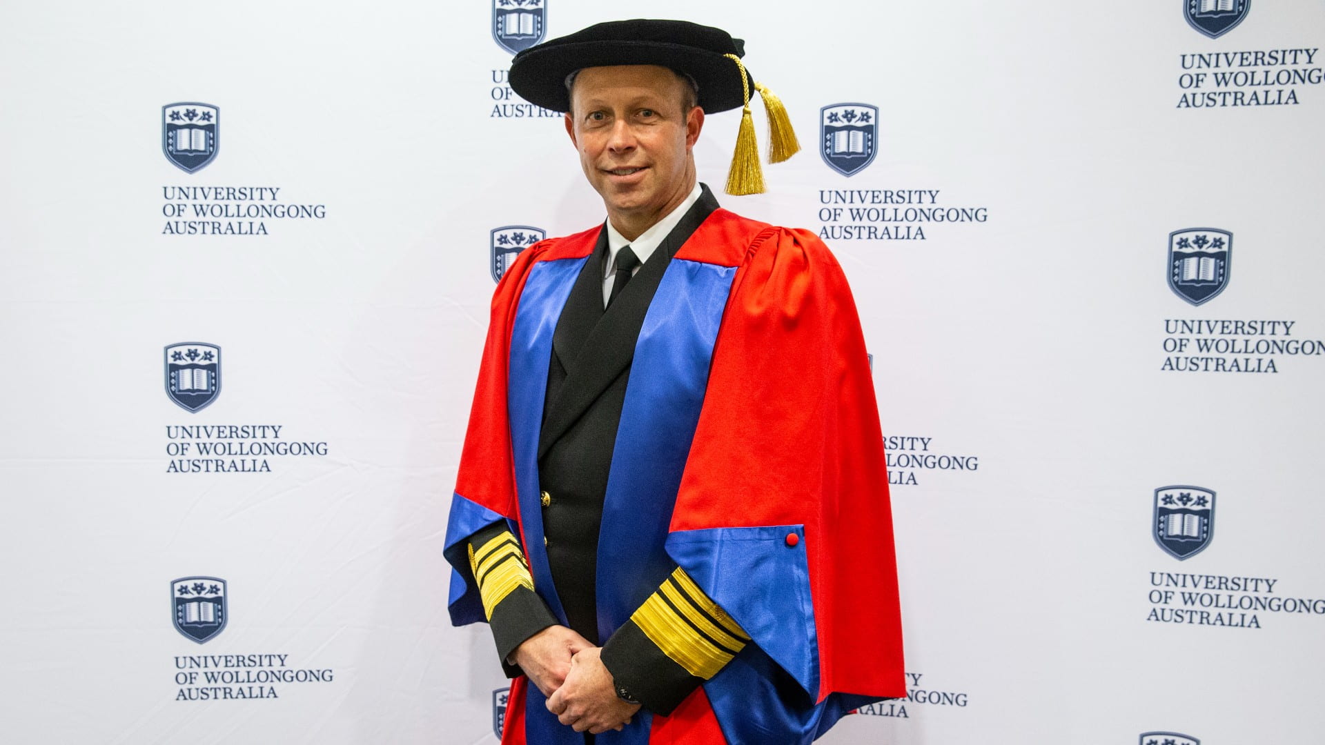 Vice Admiral Michael Noonan, wearing a red graduation gown, in front of a UOW media wall ahead of receiving his honorary doctorate. Photo: Paul Jones