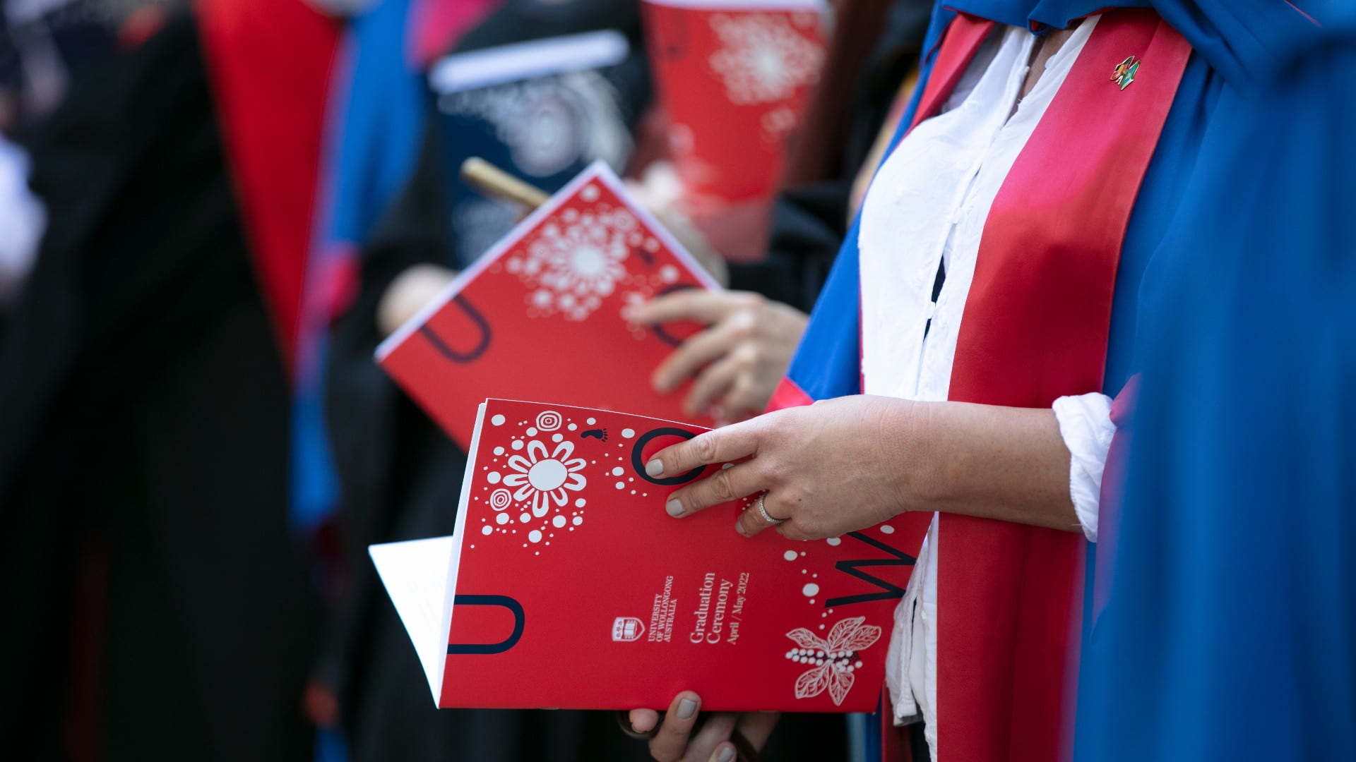 A close-up of a woman's hands, holding the bright red Graduation brochures. She is wearing a graduation robe. Photo: Paul Jones
