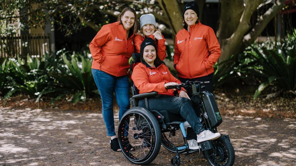 Danielle Skropeta, Tam Stutchbury, Diana King, Sharon Robinson all pose together, wearing red Homeward Bound jackets. Danielle, Tam and Sharon stand behind Diana, who is in a wheelchair. Photo: Michael Gray