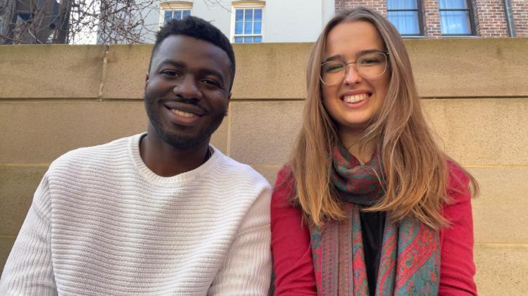 Jeremiah Edagbami and Mary Pilkinton sit side by side, each with a smile on their face. Jeremiah wears a light grey jumper while Mary wears a red jumper with a scarf. Mary has glasses.