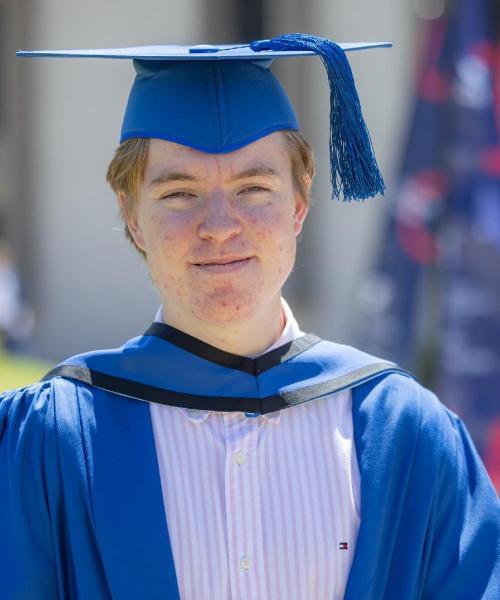Graduating student Jackson Gunn stands in front of the Sports Hub, wearing a blue cap and gown. Photo: Mark Newsham