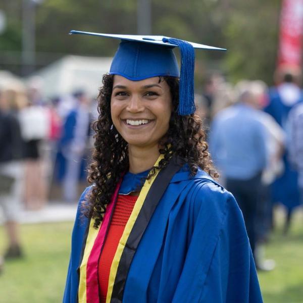 Janaya Pender, wearing a red dress, blue and red graduation gown, and blue cap, smiles at the camera. There are people gathered in the background. Photo: Mark Newsham