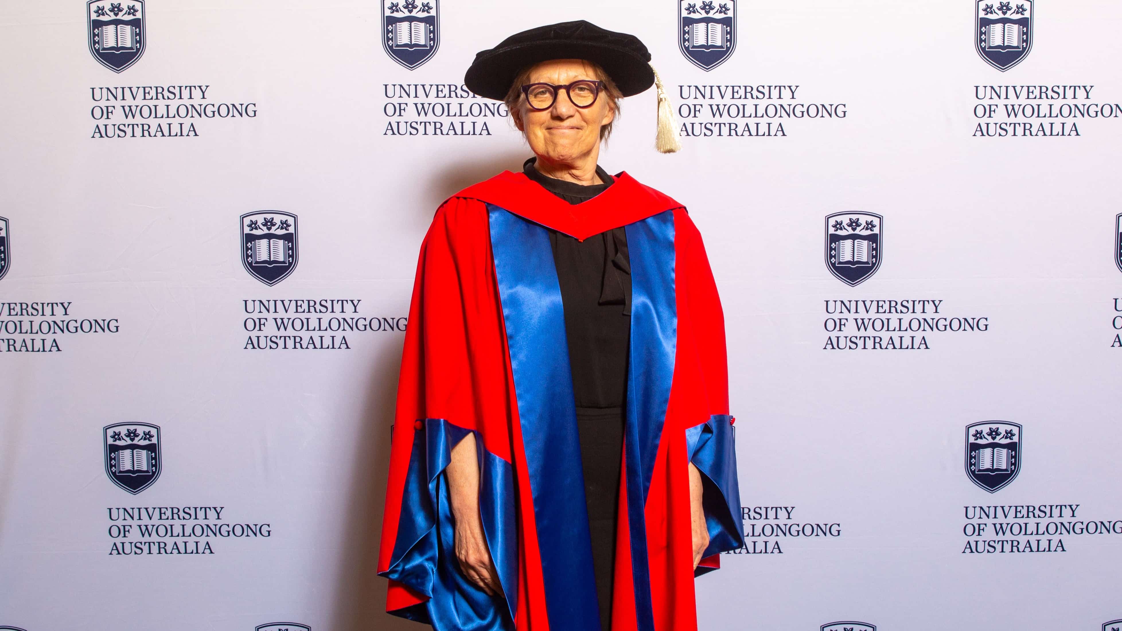 Professor Judy Raper wears a black graduation cap and red gown and stands in front of a UOW wall. Photo: Andy Zakeli