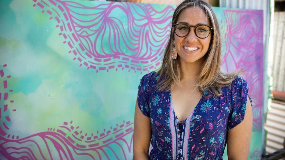 Kirli Saunders smiling against a brightly coloured canvas in the background.