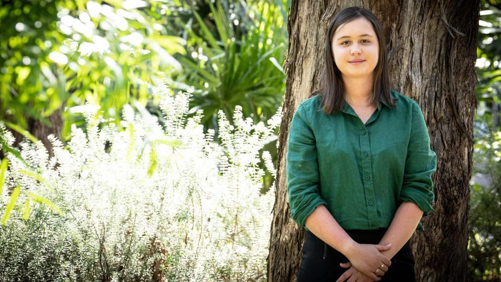 Lara Hase, wearing a green shirt, stands against a tree, with bushes in the background. Photo: Paul Jones