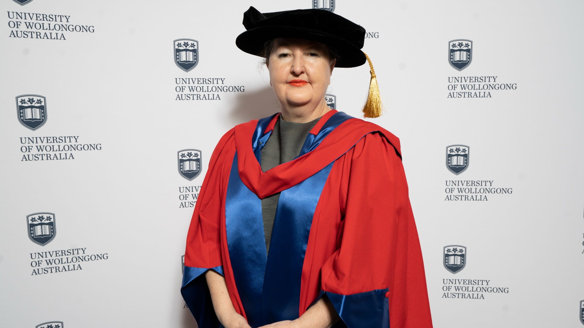 Dr Lisa Havilah, wearing a black graduation cap and blue and red graduation gown, stands in front of a UOW wall. Photo: Paul Jones
