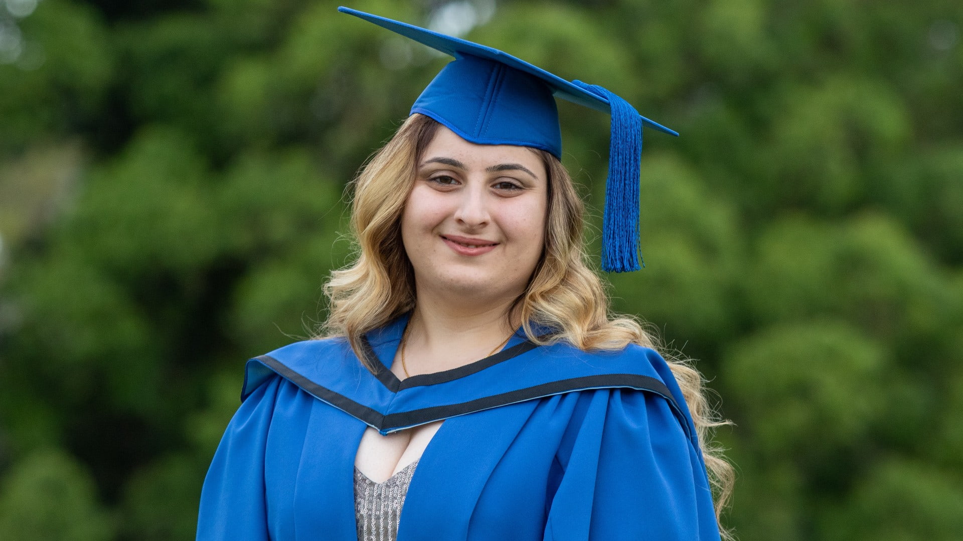 Lord Thabet, in a blue graduation gown and cap, smiles. There are trees in the background. Photo: Andy Zakeli