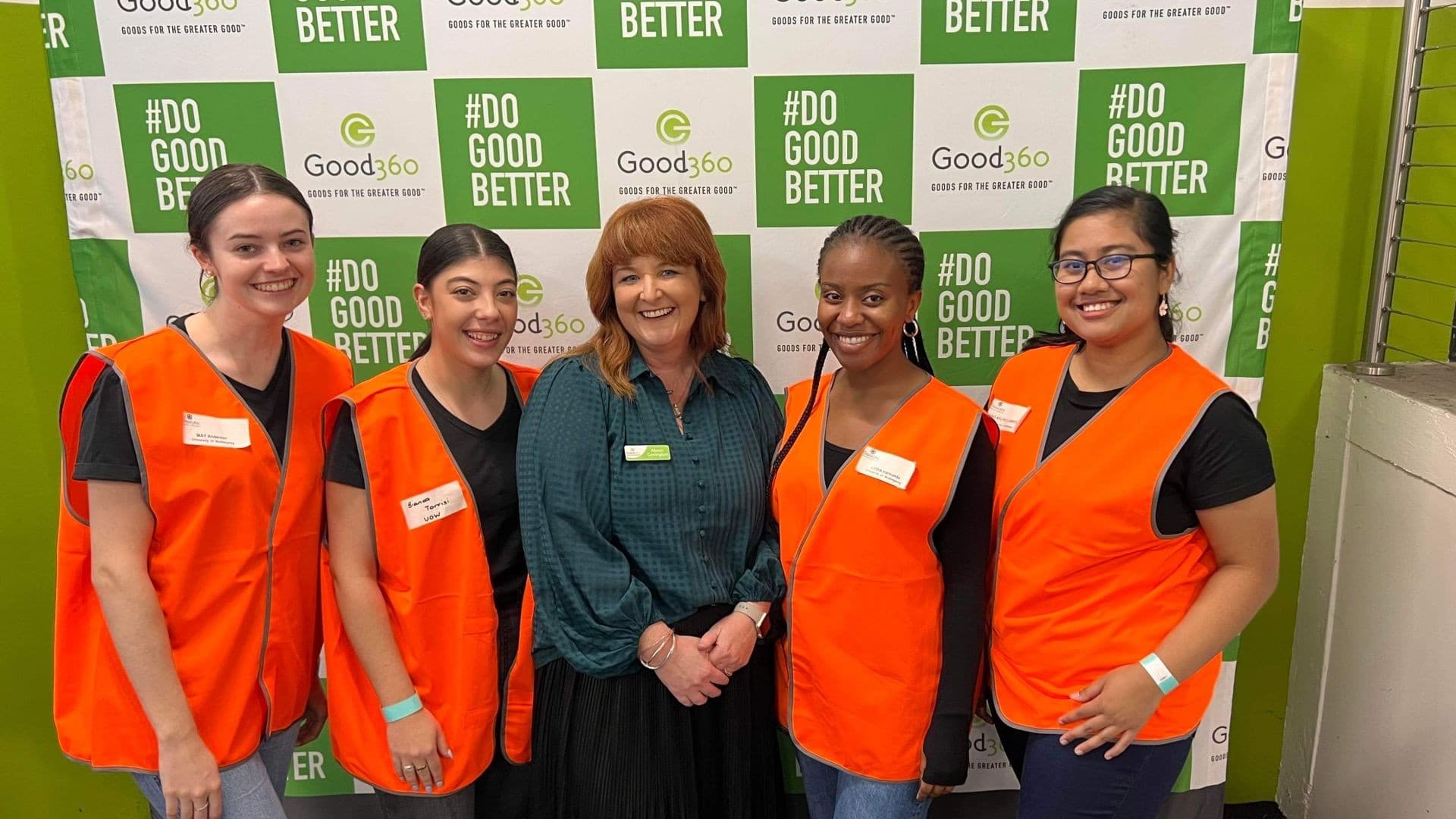 Four UOW students, wearing orange reflective vests, stand either side of Alison Covington, from Good360, who is wearing a green shirt. They are in front of a media wall. Photo: Supplied