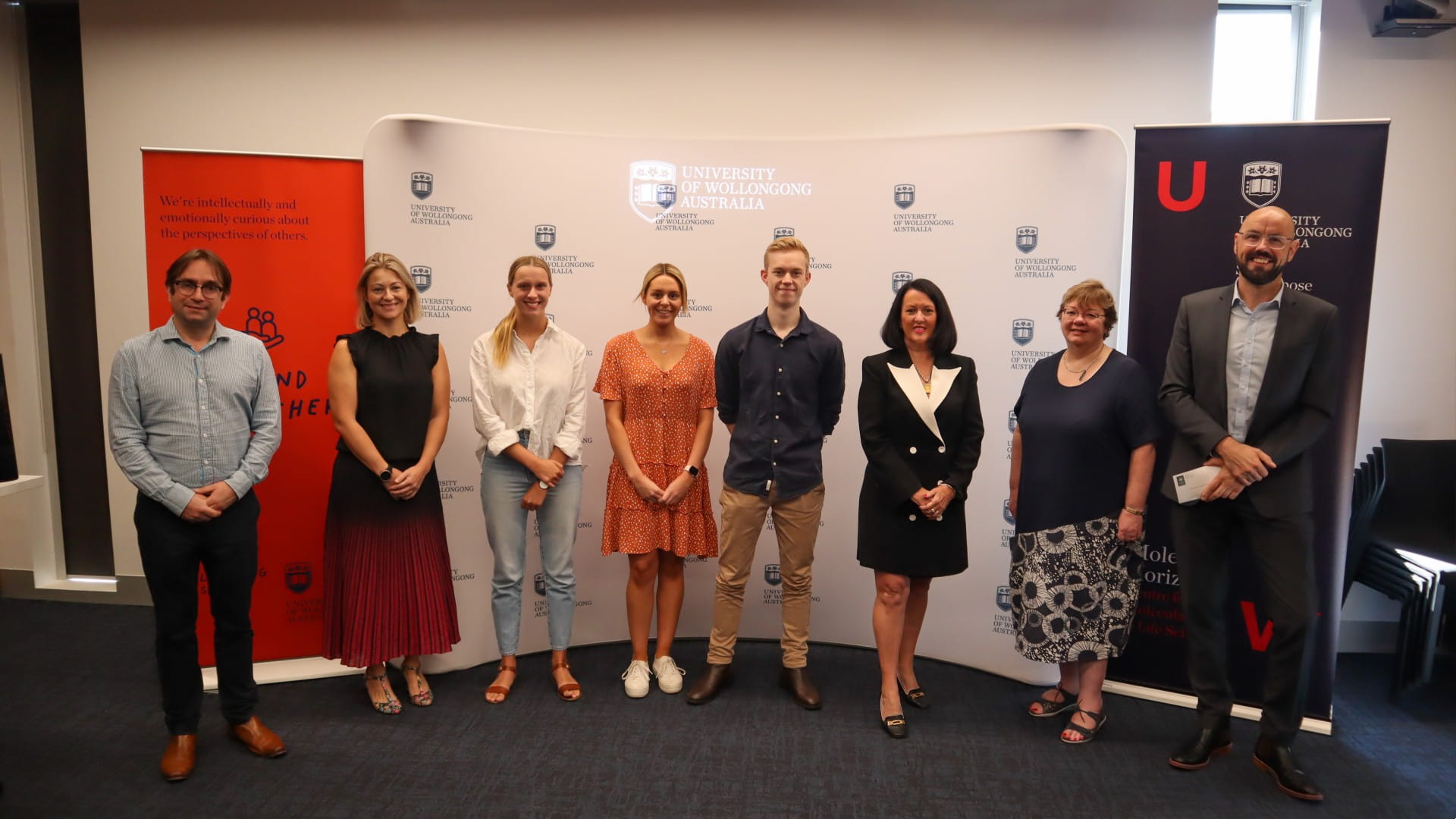 Eight people stand against a UOW backdrop during the Michael Tynan Challenge Presentation 2022.