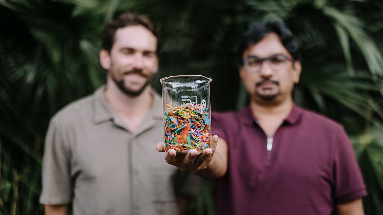 UOW PhD candidate Michael Staplevan and Professor Faisal Hai (out of focus) holding a glass beaker full of colourful plastic pieces (in focus). Photo: Michael Gray