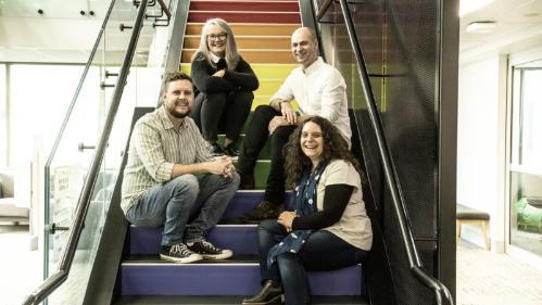 Four people sit on rainbow stairs smiling at the camera