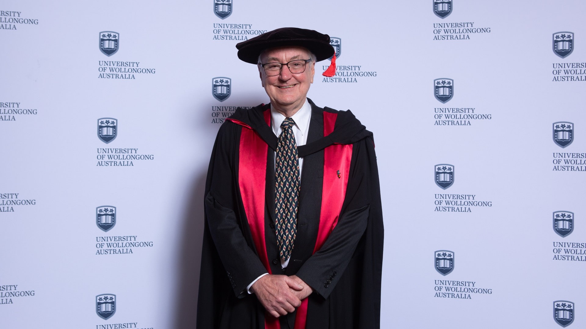 Professor Nicholas Dixon stands in front of a UOW media wall and wears a black and red graduation gown. Photo: Andy Zakeli