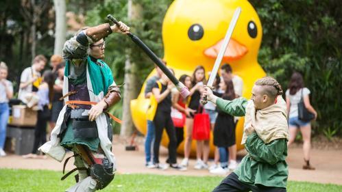 Two students take part in a pretend sword fight while a group of students watch on. The big UOW duck is in the background. Photo: Paul Jones