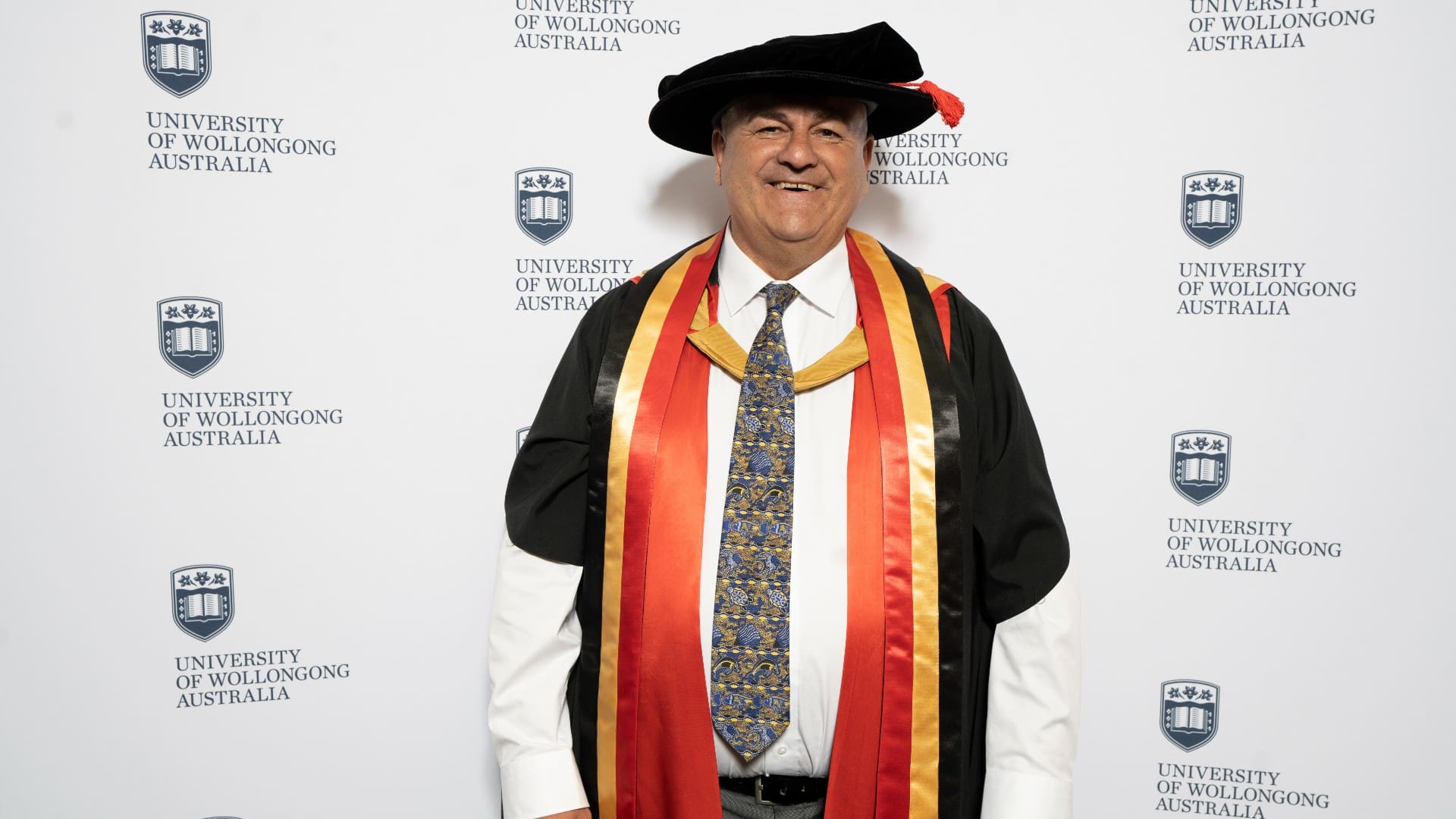 Professor Paul Chandler smiles at the camera. He wears a black graduation gown with the red, yellow and black of the Indigenous colours drapped around his neck. Photo: Paul Jones