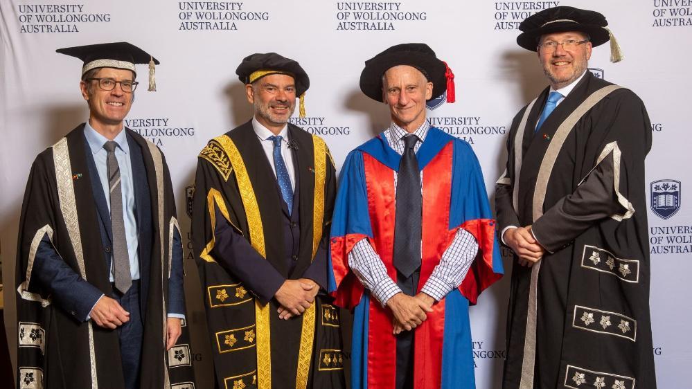 Professor Theo Farrell, Warwick Shanks, Professor Paul Else, and Professor David Currow all wear graduation gowns and caps and stand in front of a white UOW wall. Photo: Andy Zakeli
