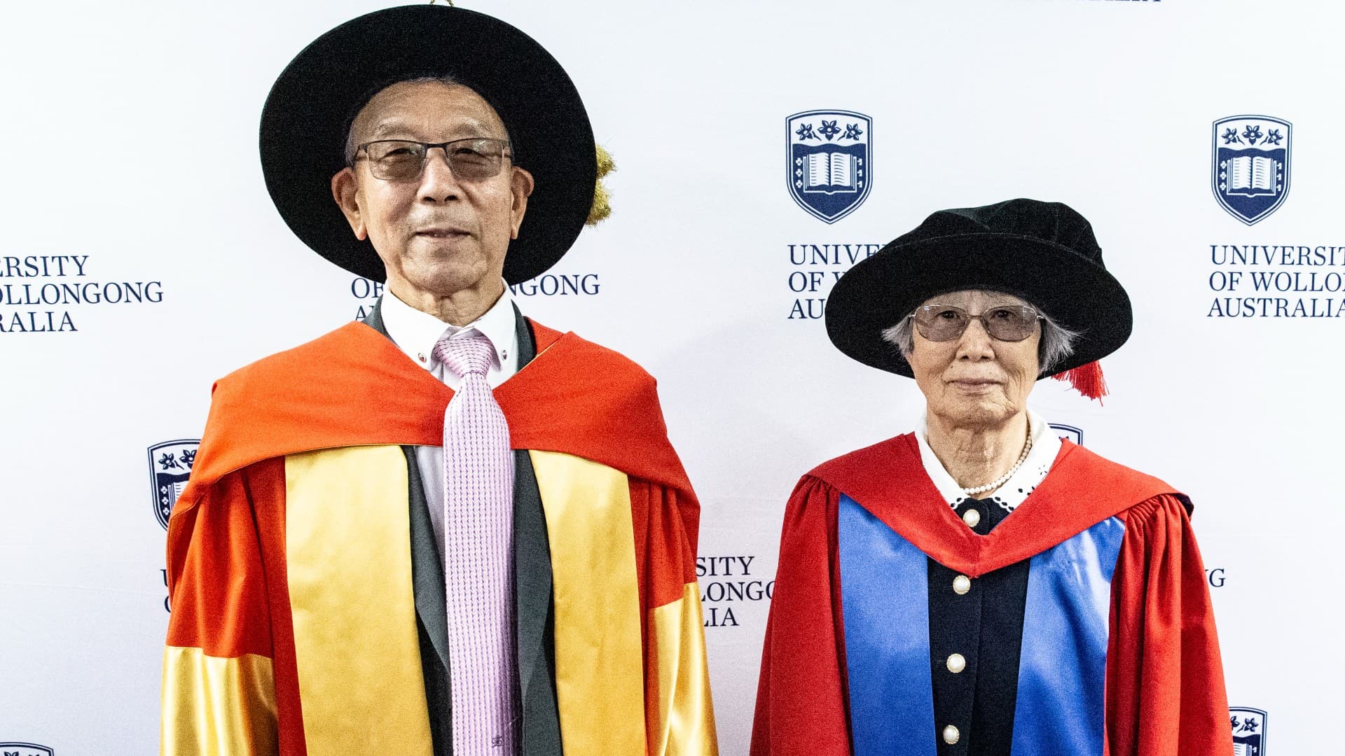 Professor Shi Xue Dou and Professor Hua Kin Liu are pictured in their graduation gowns and caps, against a UOW wall. Photo: Paul Jones