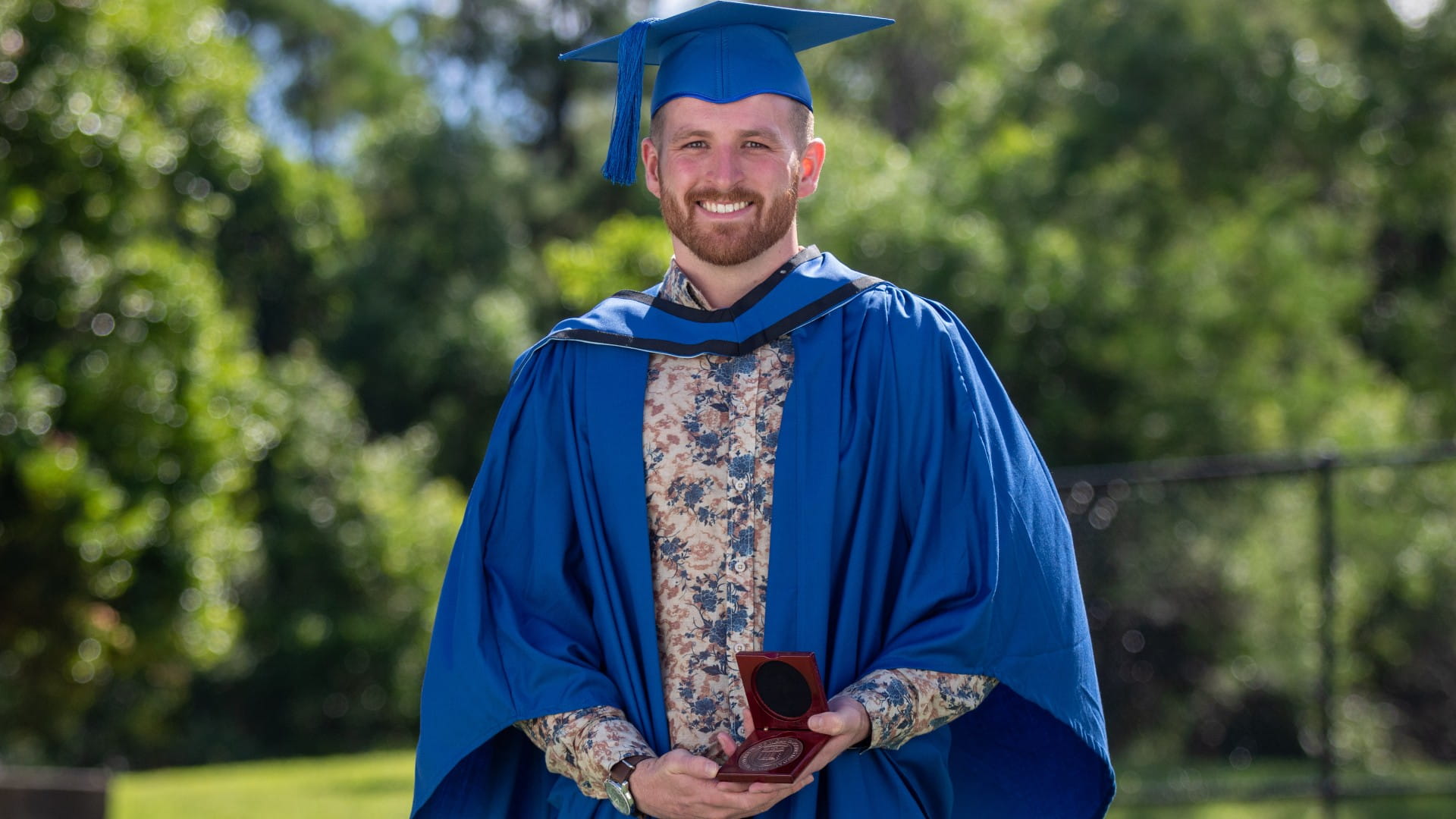 Rhys Smith, wearing a blue graduation cap and gown, holds the University medal and smiles. Photo: Andy Zakeli