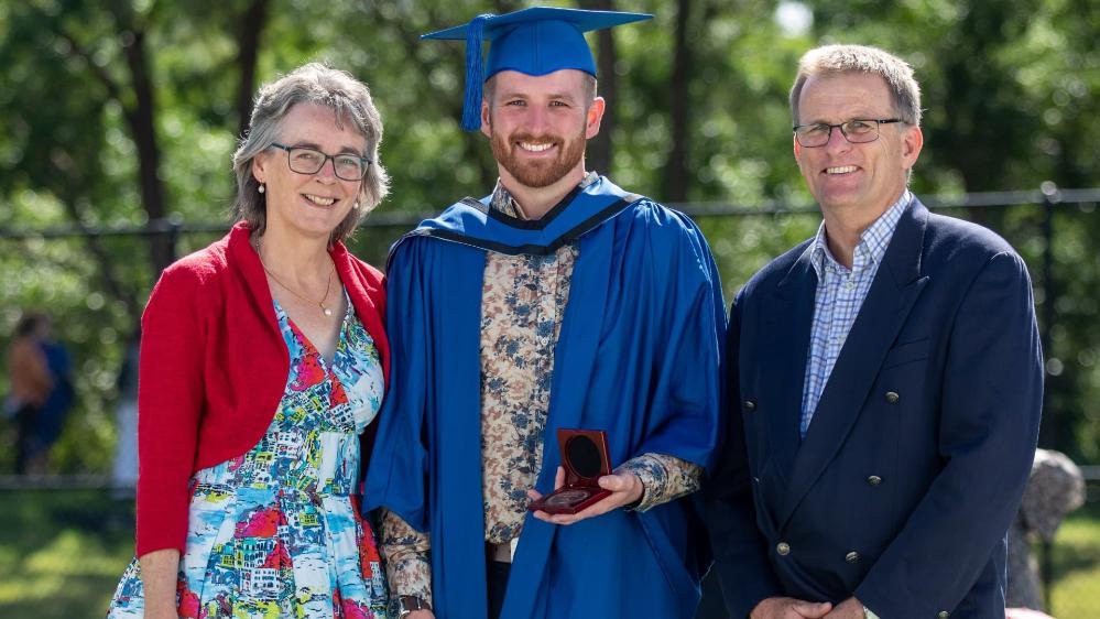 Rhys Smith (centre), wears a blue graduation cap and gown and holds the University Medal. To his left is his mother. She wears a dress and red cardigan and smiles at the camera. To his right is his father. He wears a blue suit and smiles at the camera. Photo: Andy Zakeli