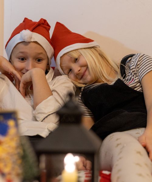 Two children sit together on a sofa and they are wearing Santa hats
