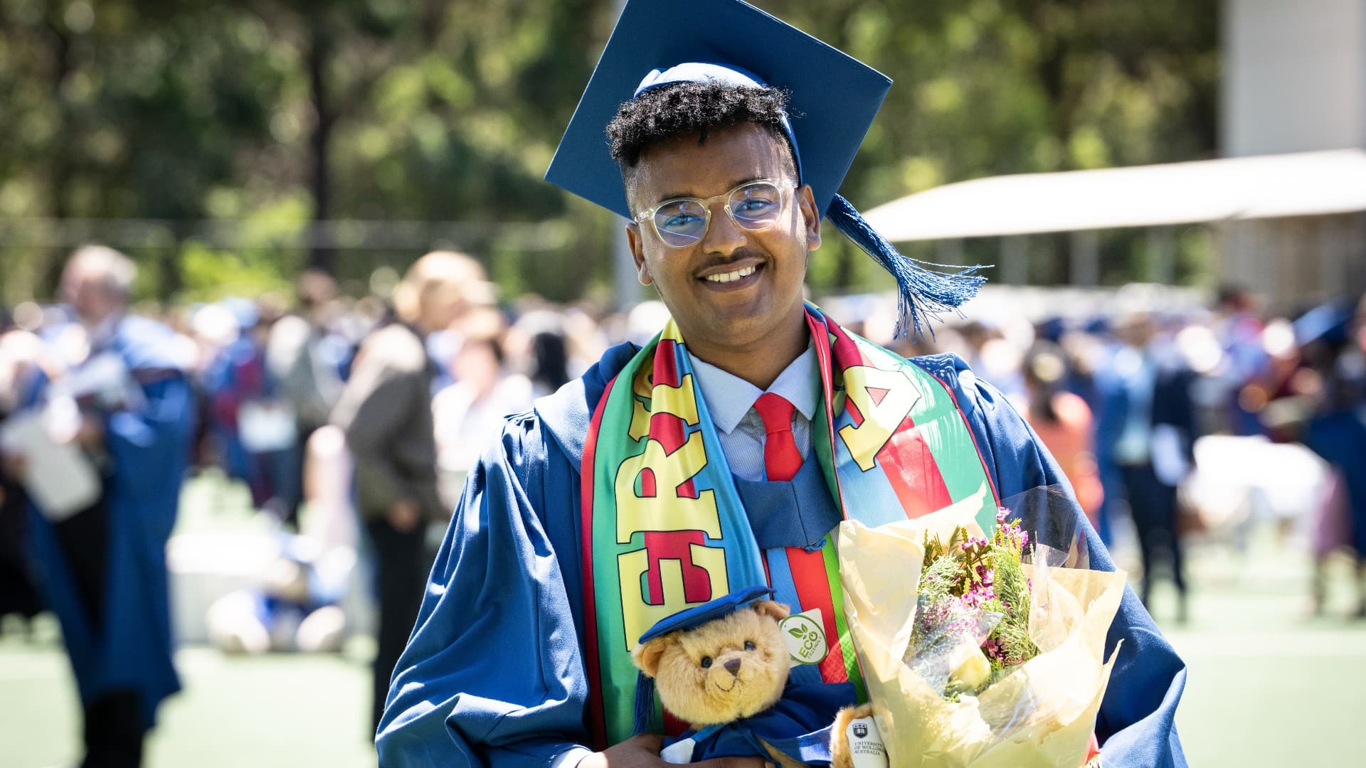 Teklemariam Mengistu wears the blue UOW graduation gown and cap, with a flag of Eritrea drapped around his neck. He holds a teddy bear and bunch of flowers and is smiling at the camera. Photo: Paul Jones