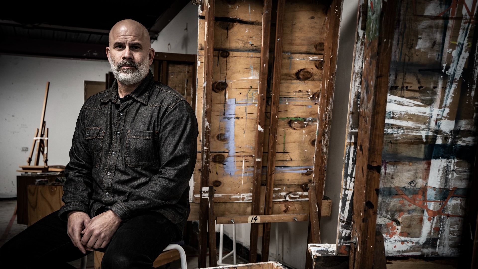 Teo Treloar, wearing a black jacket and pants, sits on a stool in an art studio, surrounded by paint on the walls. Photo: Paul Jones