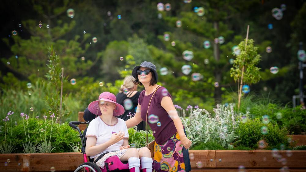 A woman in a wheelchair and a woman standing next to her look at the new playground while bubbles float in the air. Photo: Paul Jones
