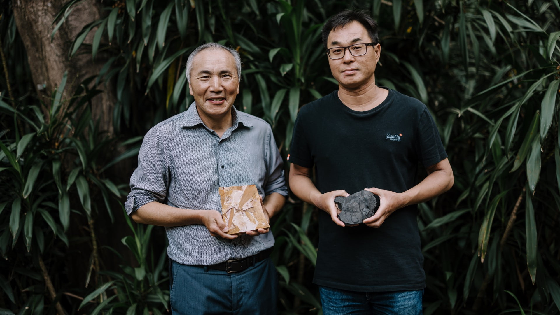 Professor Guang Shi and Dr Sam Lee stand in front of a green bush each holding a rock sample. Photo: Michael Gray