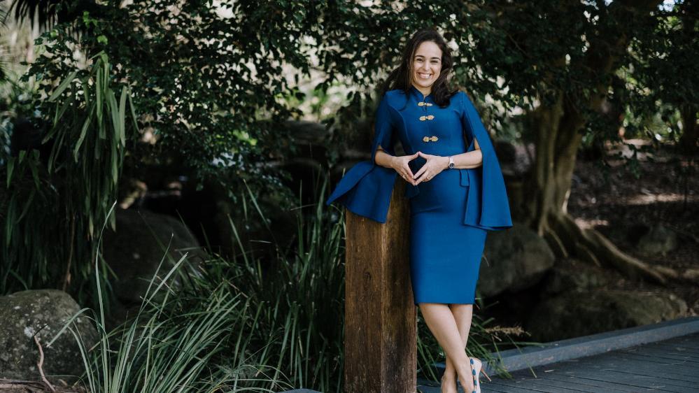 Izabela Pereira Watts wears a blue dress against a leafy background. She is leaning against a pole. Photo: Michael Gray