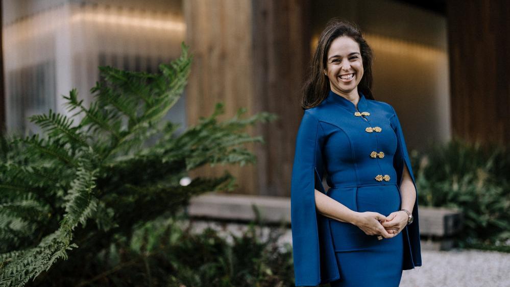 Dr Izabela Pereira Watts, wearing a blue dress with gold buttons, stands in front of a building smiling at the camera.