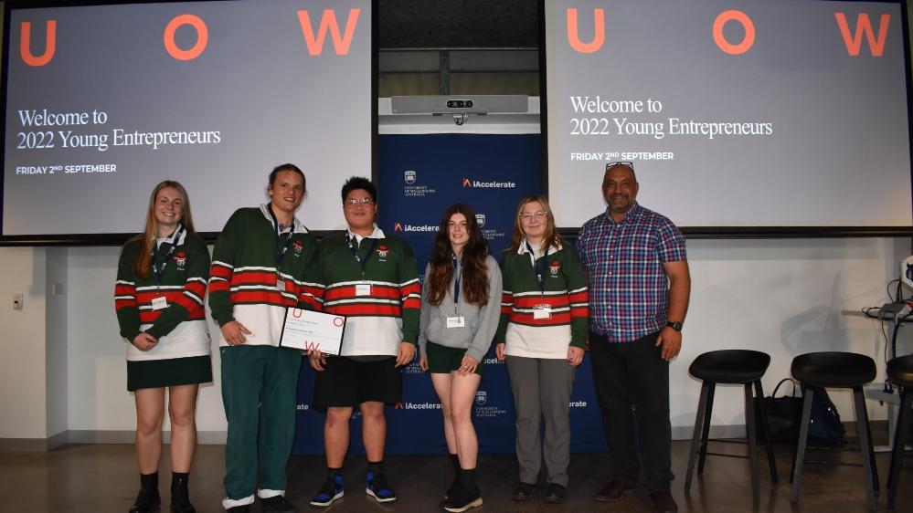Five students from Bomaderry High School, all wearing green, red and white jumpers, stand in front of two screens with UOW on them.