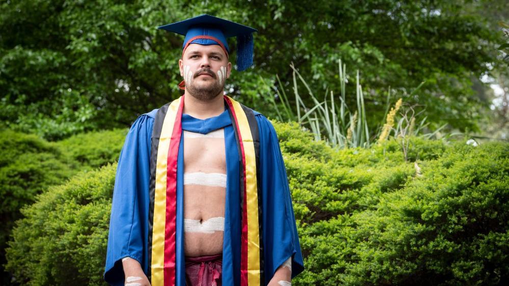 UOW graduate Zachary Stewart, wearing graduation gowns with the Indigenous colours and his traditional dress, stands against a green bush background. Photo: Paul Jones