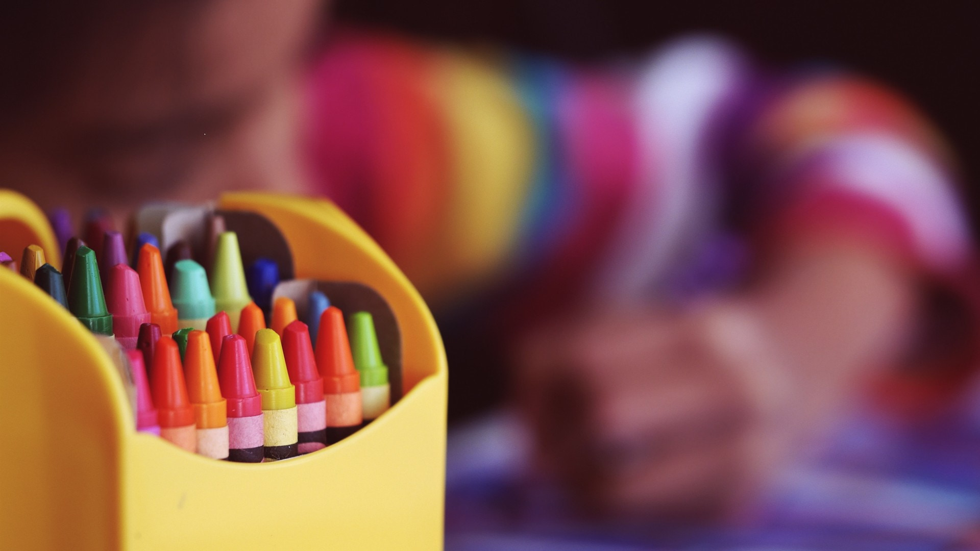An image of a colourful box of crayons, with a child seen drawing in the background. Photo: Aaron Burden/Unsplash