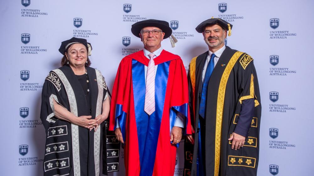 Professor Joe Chicharo, wearing a blue and red graduation gown and cap, stands between Vice-Chancellor Professor Patricia Davidson, in a black gown, and Deputy Chancellor Rob Ryan, in a black gown. Photo: Andy Zakeli