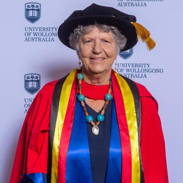 Aunty Joyce Donovan is smiling and standing in front of a UOW media wall wearing a graduation gown and cap. Photo: Andy Zakeli