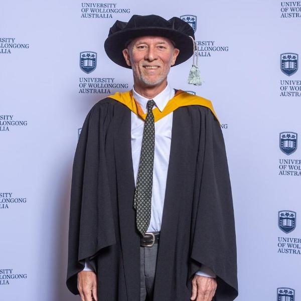Professor Frank Deane in a black and gold gown and cap in front of a UOW media wall. Photo: Andy Zakeli
