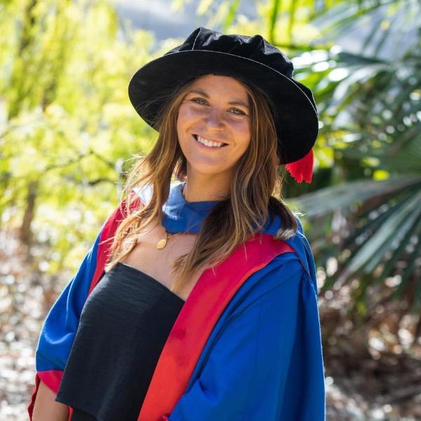Dr Freya Croft, wearing a black dress, blue and red graduation gown, and black cap, smiles at the camera. There are green bushes in the background. Photo: Andy Zakeli