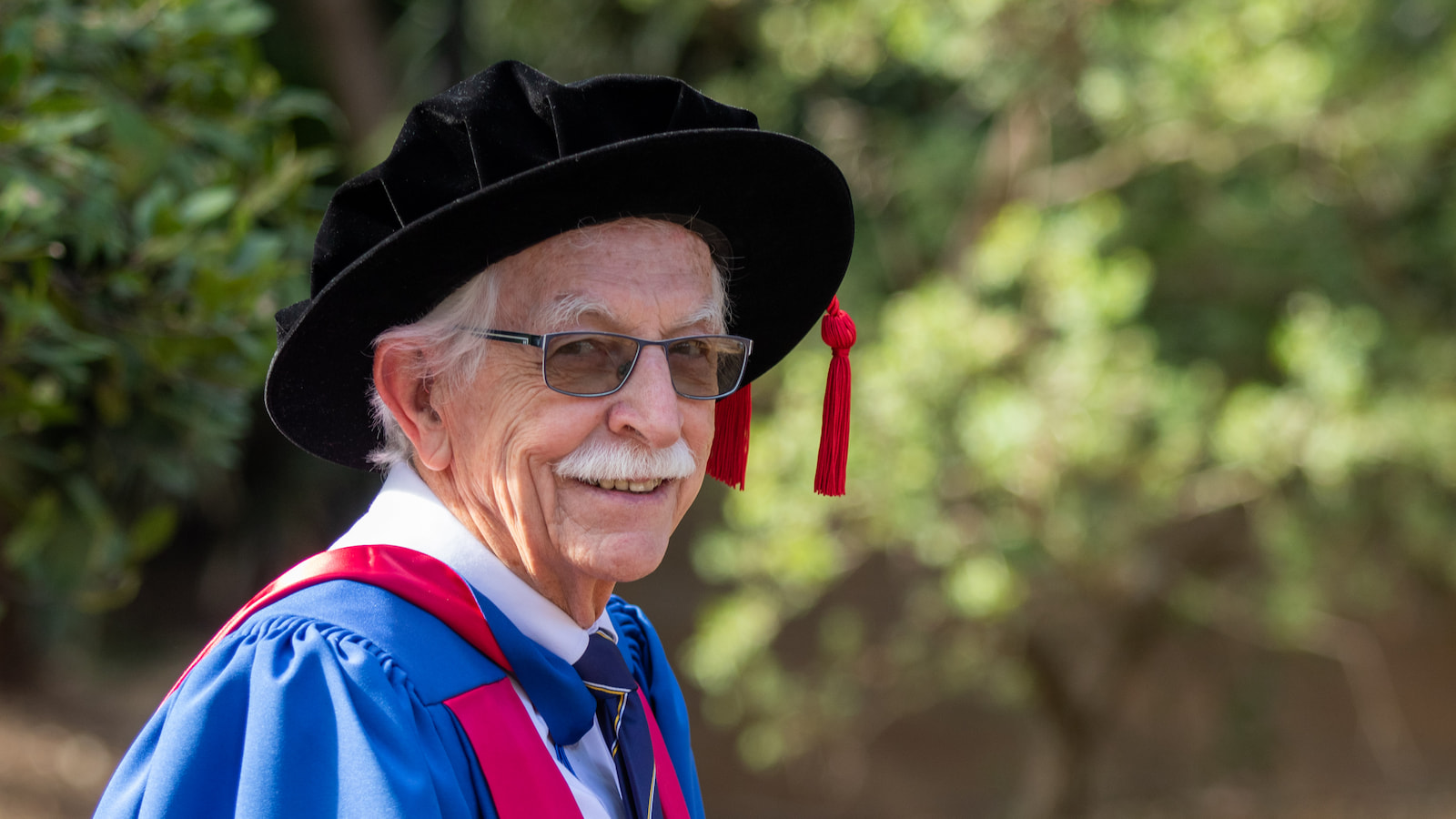 Dr Leigh Robinson, wearing a black PhD cap and blue and red gown, looks at the camera. Photo: Andy Zakeli