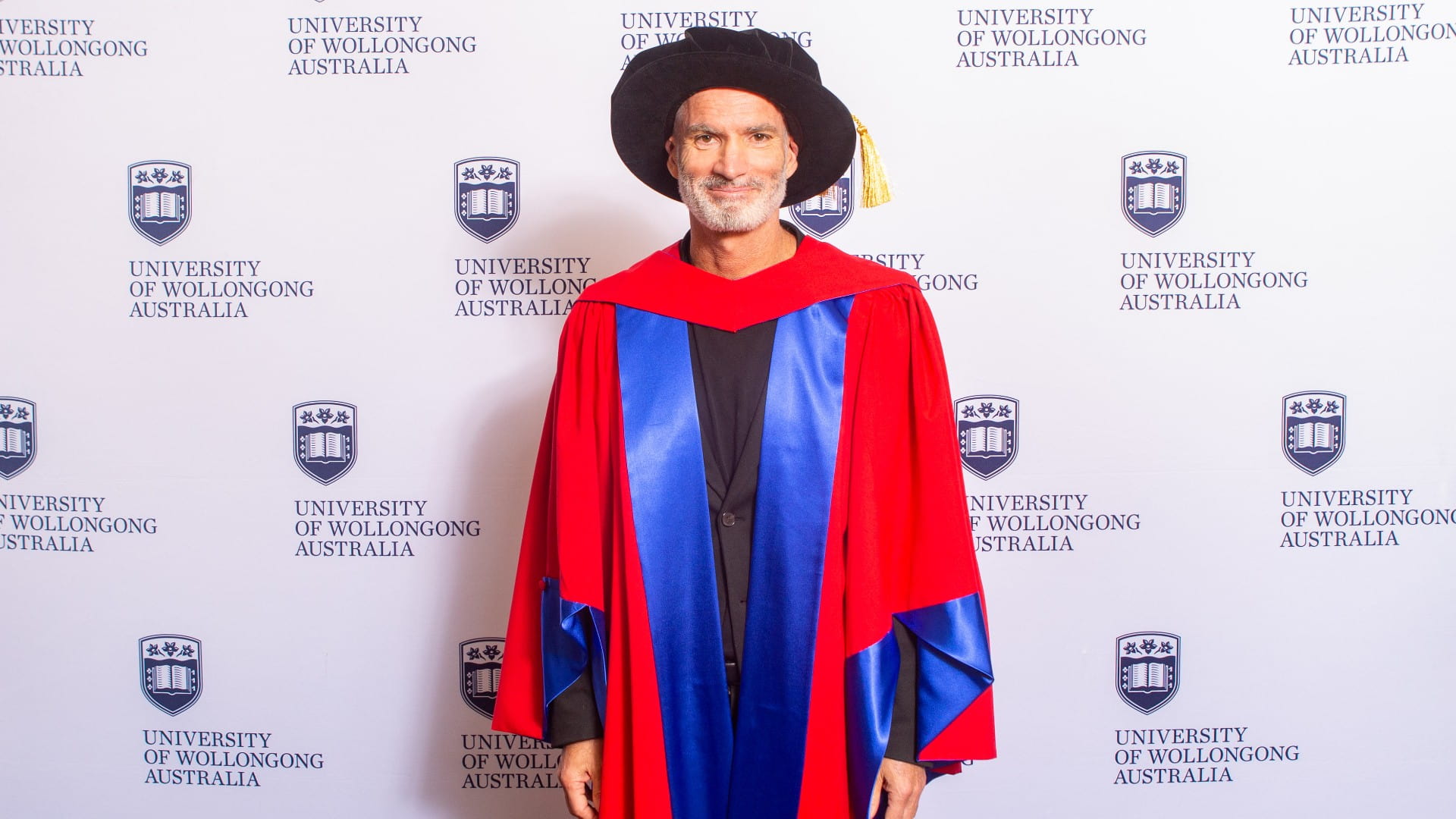 Craig Foster wears a graduation gown and cap and stands in front of a UOW media wall. Photo: Andy Zakeli