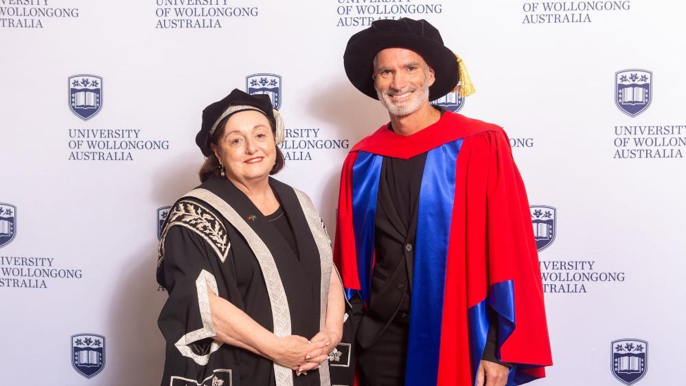 UOW Vice Chancellor Professor Patricia M Davidson and Craig Foster both wear a graduation gown and cap and stands in front of a UOW media wall. Photo: Andy Zakeli