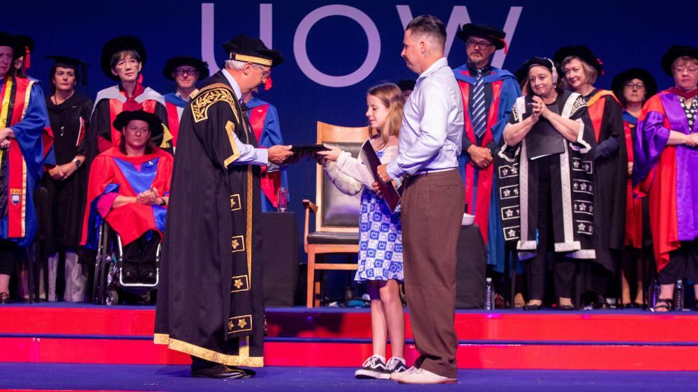 Ben and Grace Livingstone stand on the graduation stage accepting a PhD cap from UOW Chancellor Michael Still, while a crowd in the background looks on. Photo: Andy Zakeli