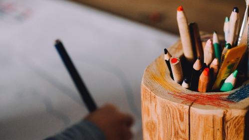 A pot of pencils is seen in the foreground with a child writing in the background. Photo: Marcus Spiske/Unsplash