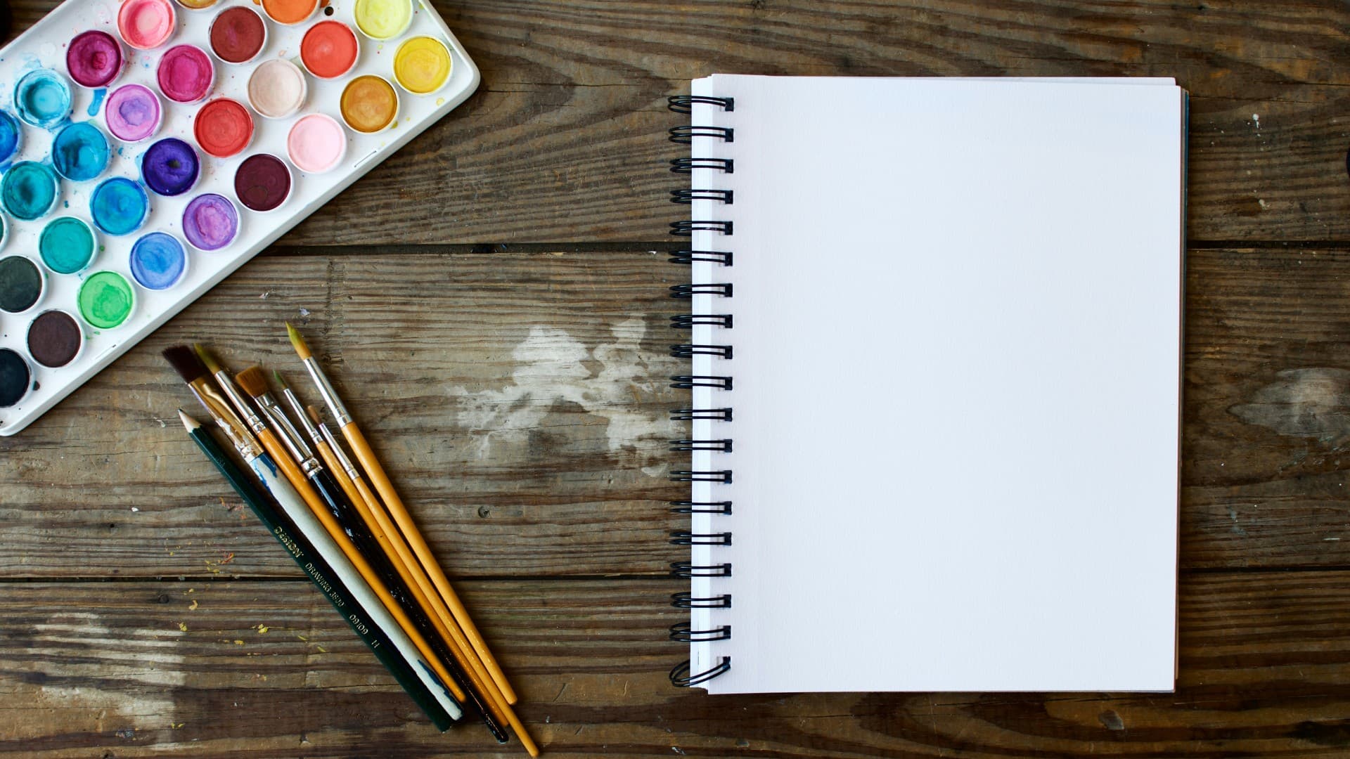Watercolour paints, paintbrushes and a notebook are laid on a table. Photo: Unsplash
