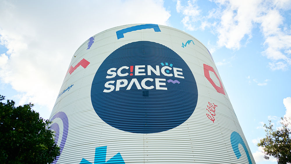 A large cylindrical building with a logo reading Science Space in a dark circle on its facade. The building is decorated with colorful geometric shapes against a light background. The sky is partly cloudy.