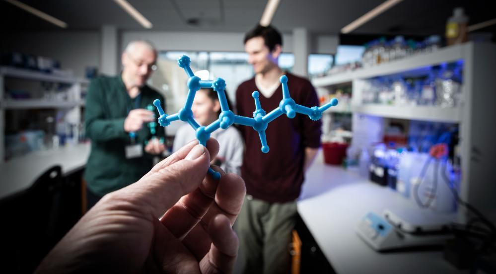Associate Professor Aaron Oakley holds an example of the 3D chemistry model. Next to him is student Karlee Symonds and Dr Patrick McClosker, who helps Karlee in the lab. They are situated in a laboratory and are all smiling. In the foreground, a hand holds a blue 3D printed chemistry model.  Photo: Paul Jones