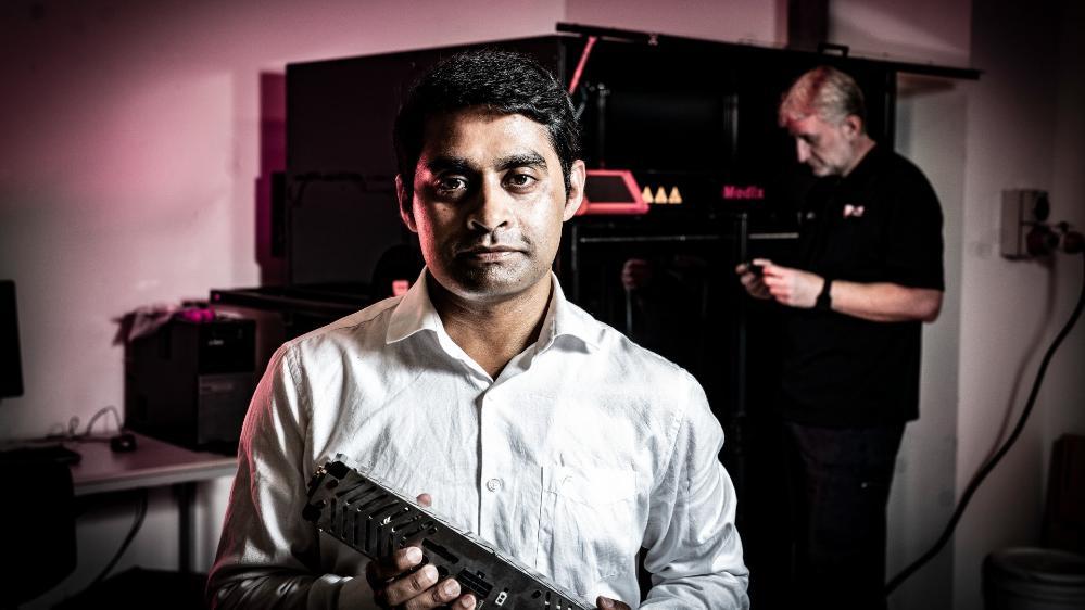 Dr Aziz Ahmed stands in the foreground, holding a piece of engineering equipment. He wears a white shirt and has a smile on his face. Photo: Paul Jones