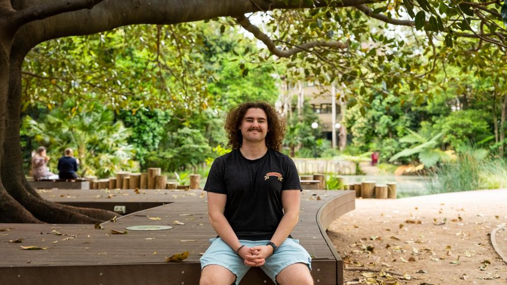 UOW student Luke Bradbery, wearing a black shirt and blue shorts, sits with the Wollongong campus in the background. Photo: Adam Mansfield