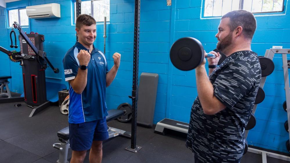 UOW College student Patrick Mitchell helps out a client in the gym, who is lifting a barbell. Patrick wears a blue shirt and is pretending to lift weights. Photo: Mark Newsham