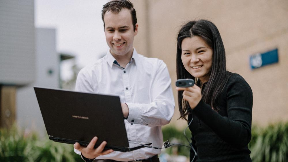 Researchers Joshua Thompson and Yunjia Lei stand next to each other. Joshua is looking at a laptop while Yunjia is holding a device. Photo: Michael Grey