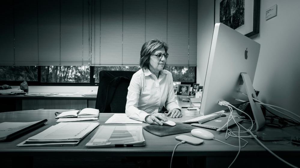 Black and white image. Professor Judy Raper sits at her computer in her office at UOW. She wears glasses and is looking at the computer.